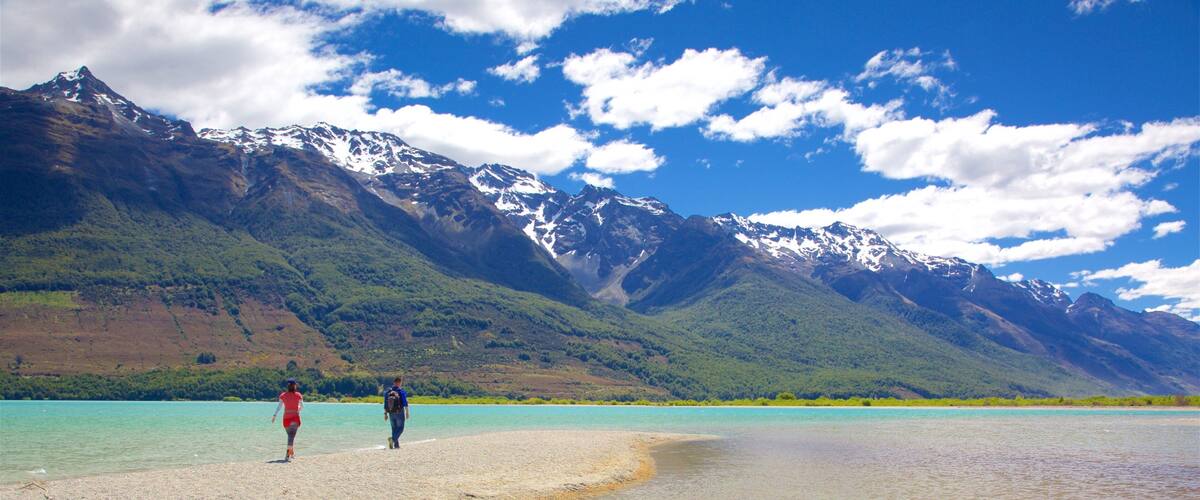 A beach with turquoise water, people walking, and snow-capped mountains in the background.