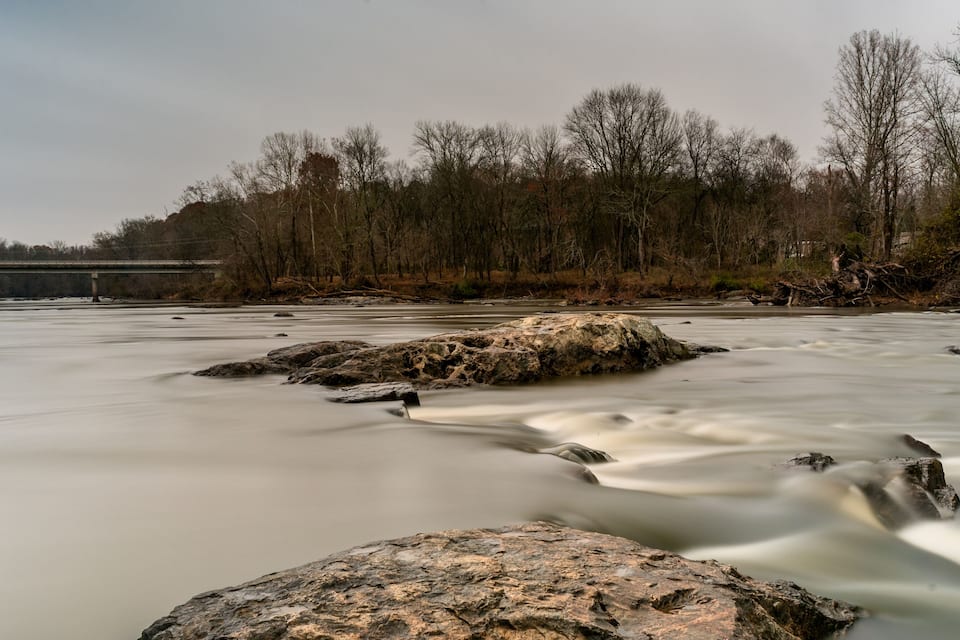 Une rivière à l’eau qui coule paisiblement, avec des rochers et un pont en arrière-plan.