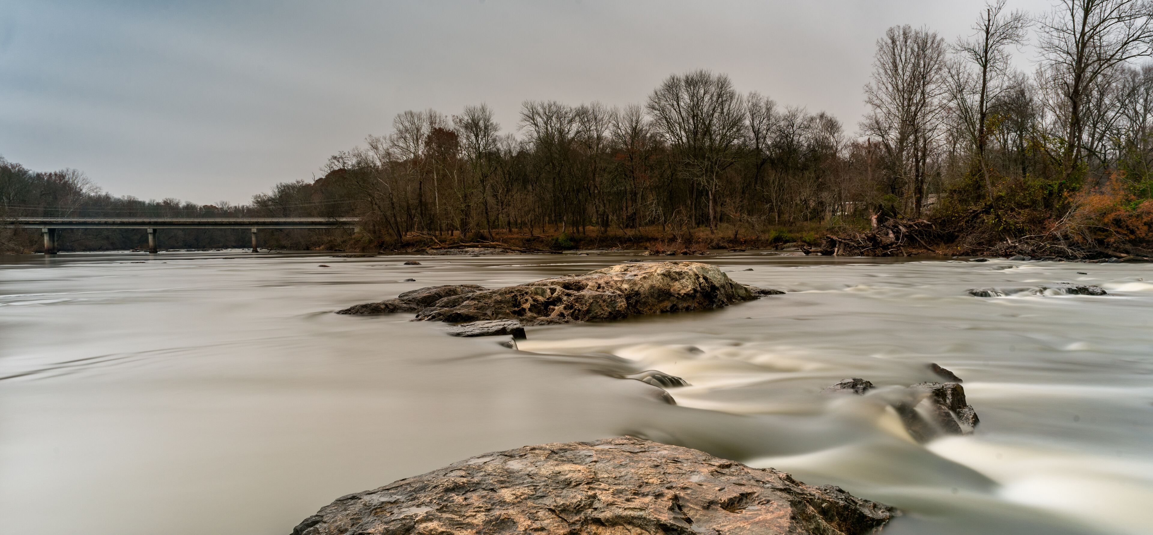 A river with smooth water flow, rocks, and a bridge in the background.