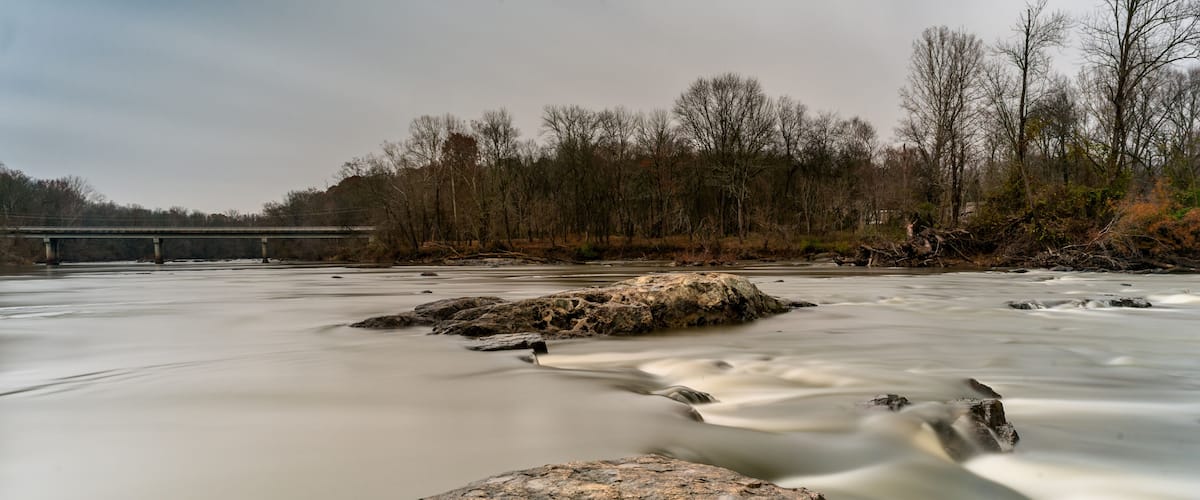 A river with smooth water flow, rocks, and a bridge in the background.
