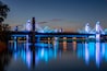 Un pont illuminé de lumières bleues dont le reflet se mire sur l’eau au crépuscule.