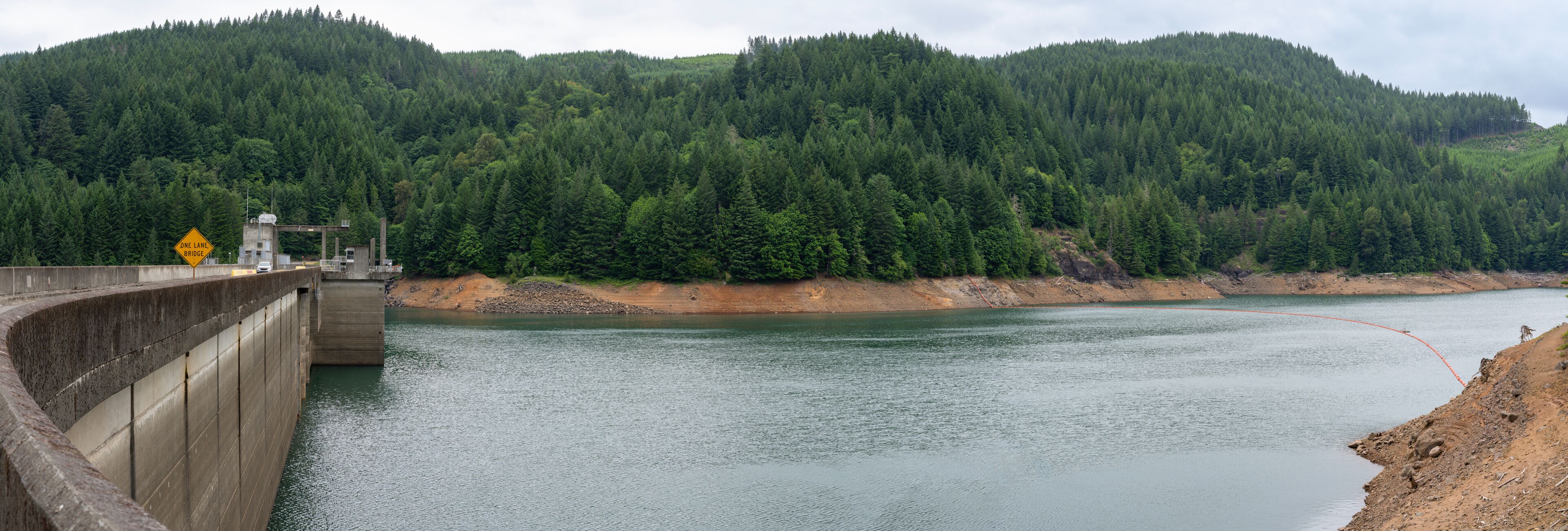 A dam with a one-lane bridge, surrounded by dense forest and a river.