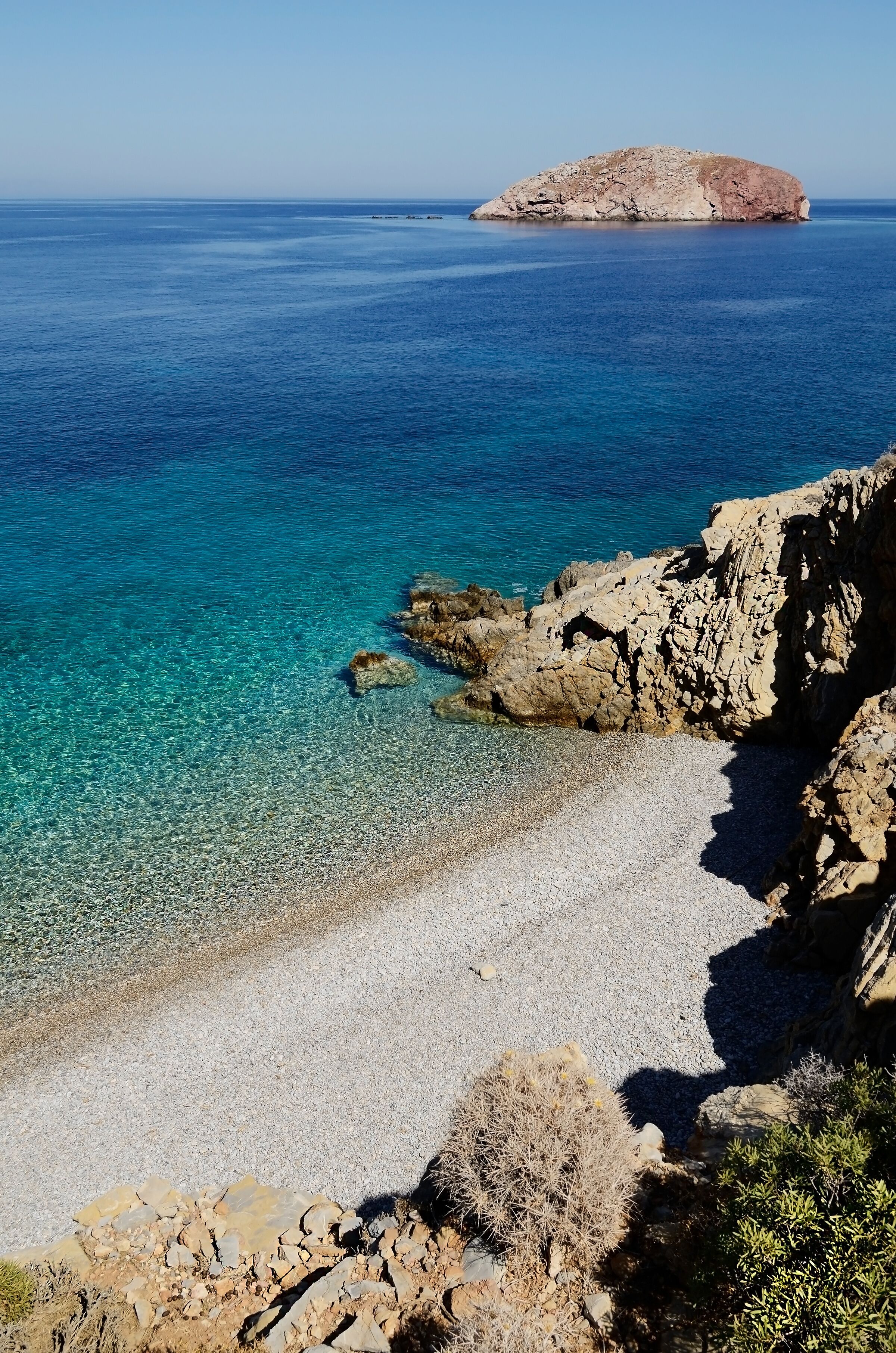 A coastal landscape with clear blue waters, a pebbly beach, rocky cliffs, and a distant island.