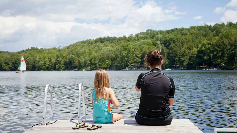 Two people sitting on a wooden dock by a lake with a sailboat in the distance and a forested hill in the background.