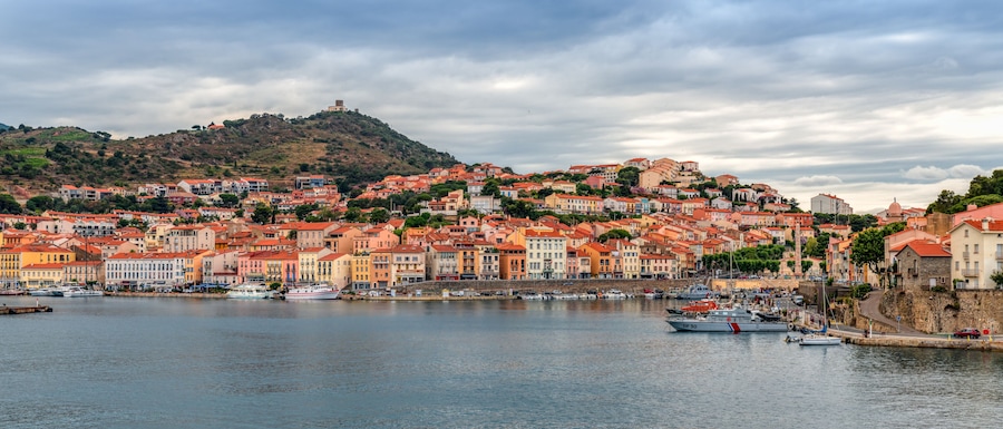Uma cidade costeira com edifícios coloridos, uma fortaleza no alto de uma colina e uma marina com barcos.