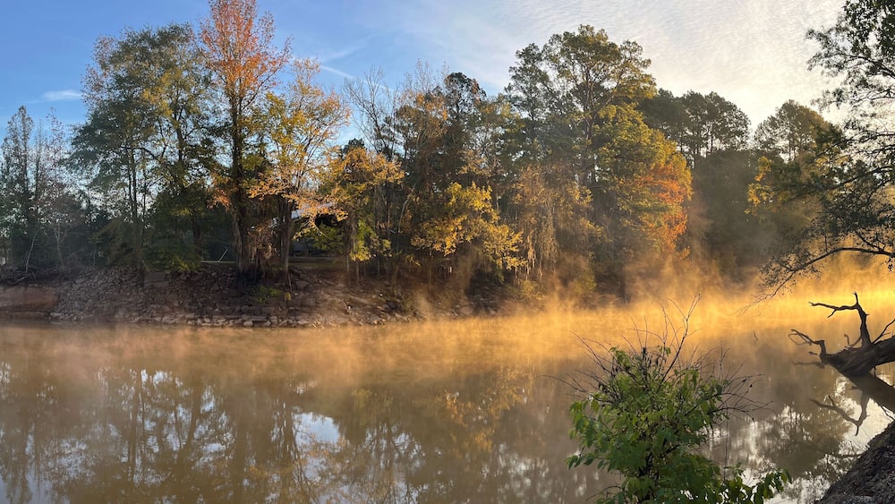 A misty river with autumn trees reflecting in the water, surrounded by a forest and a building on the left.