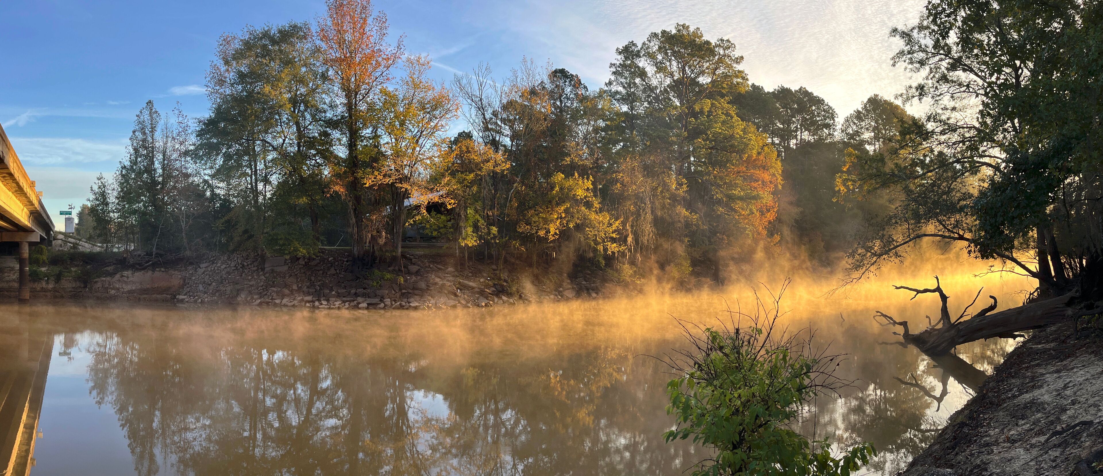 A misty river with autumn trees reflecting in the water, surrounded by a forest and a building on the left.