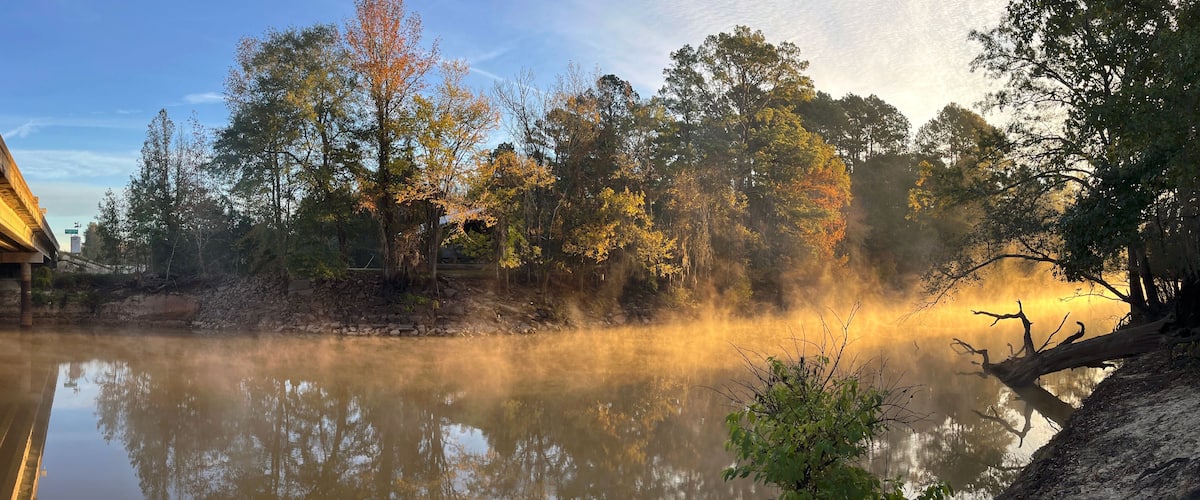 A misty river with autumn trees reflecting in the water, surrounded by a forest and a building on the left.