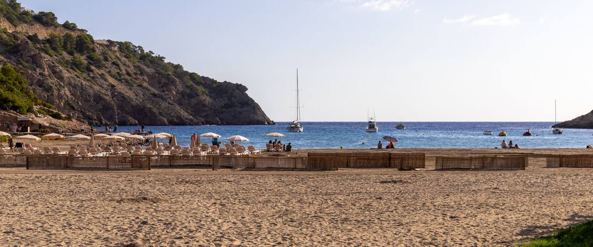 A beach with umbrellas, people, boats, and a rocky hillside.