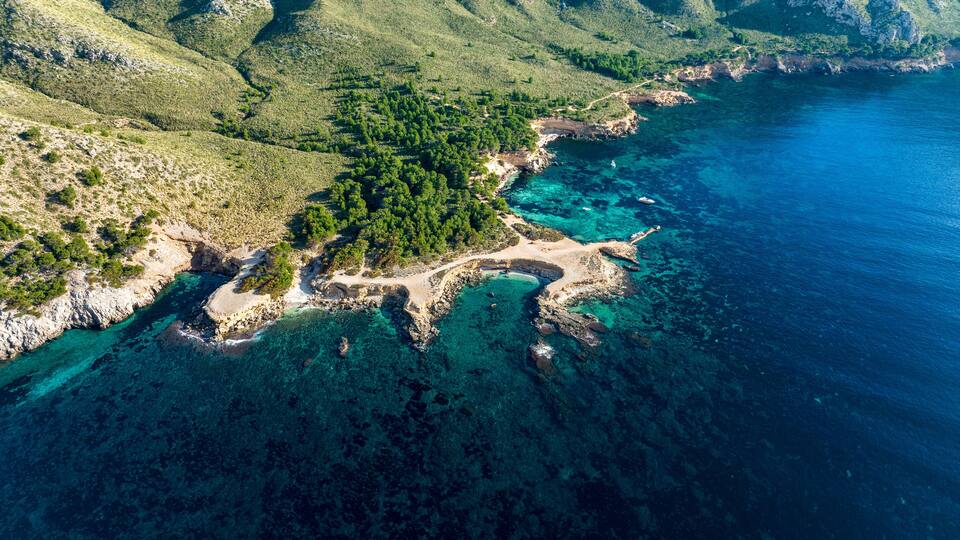 An aerial view of a coastal landscape with a rocky shoreline, clear turquoise waters, and dense green vegetation on the hills.
