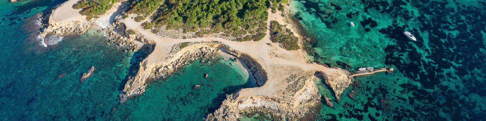 An aerial view of a coastal area with clear turquoise waters, rocky shores, and dense green forests.