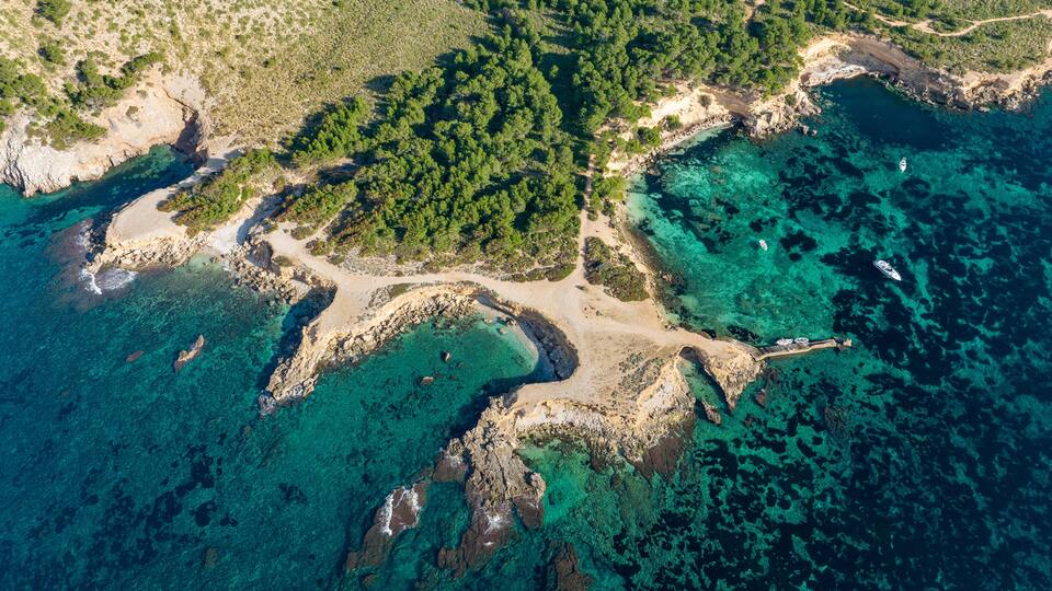 An aerial view of a coastal area with clear turquoise waters, rocky shores, and dense green forests.
