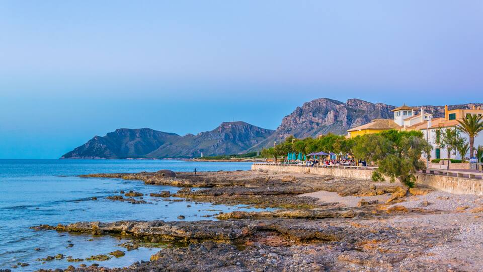 A rocky shoreline with a mountain in the background and a building with a red roof.