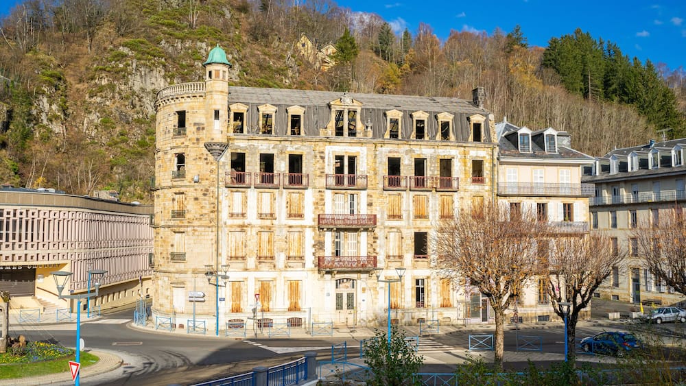 A historic stone building with multiple balconies and a turret, surrounded by a modern urban environment with a clear sky.