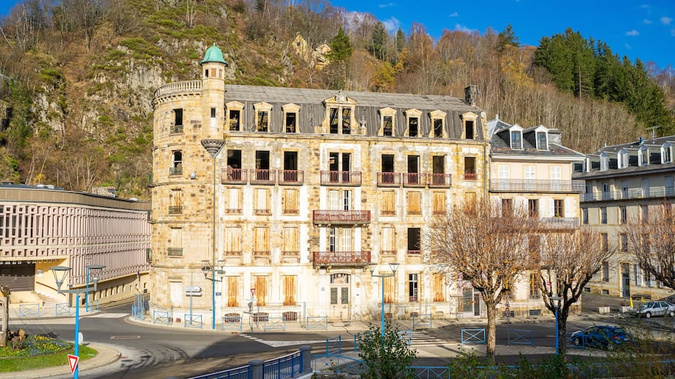 A historic stone building with multiple balconies and a turret, surrounded by a modern urban environment with a clear sky.