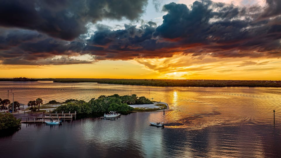 A sunset over a calm river with boats, a dock, and a small island with vegetation.
