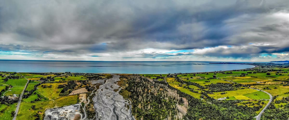 A coastal landscape with a winding road, green fields, and a large body of water under a cloudy sky.