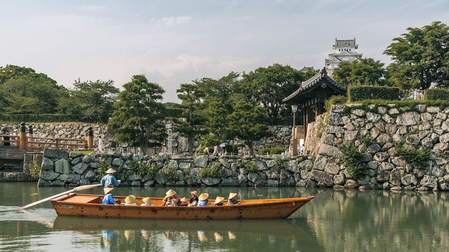 A traditional wooden boat with passengers, a stone wall, and a pagoda-like structure in the background.