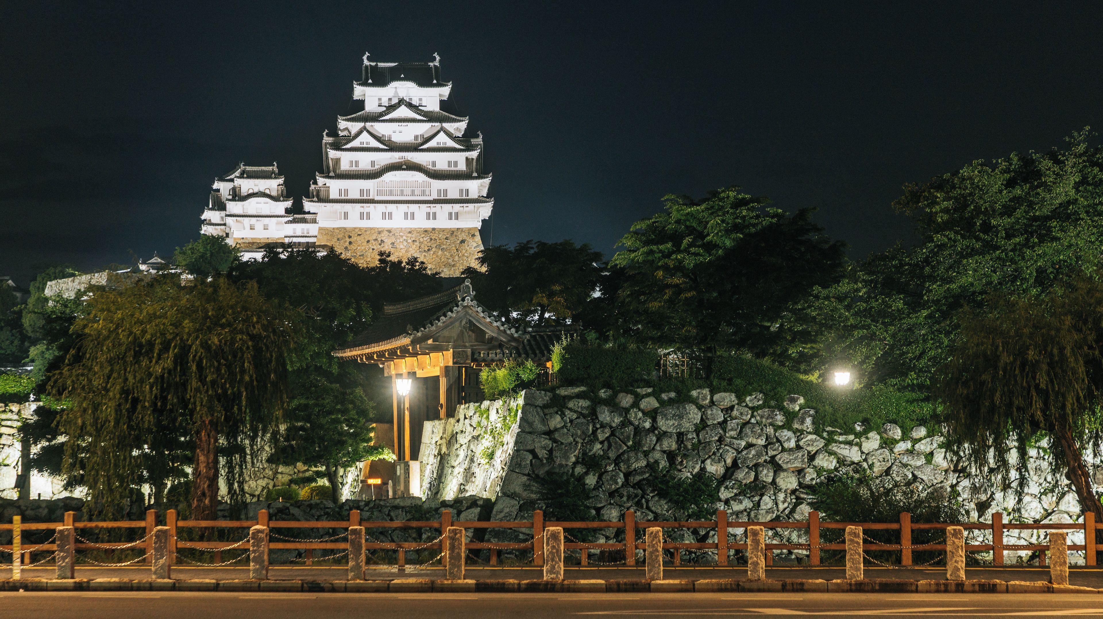 A traditional Japanese castle at night, with a stone wall and a wooden fence in the foreground.