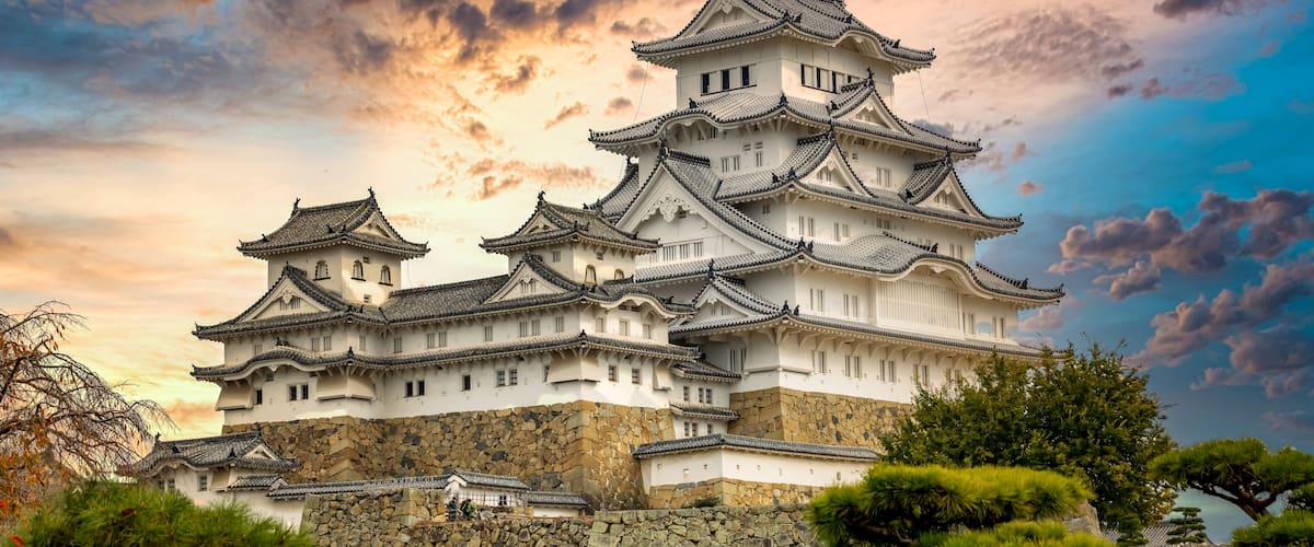 A traditional Japanese castle with multiple tiers and a stone base, surrounded by greenery.