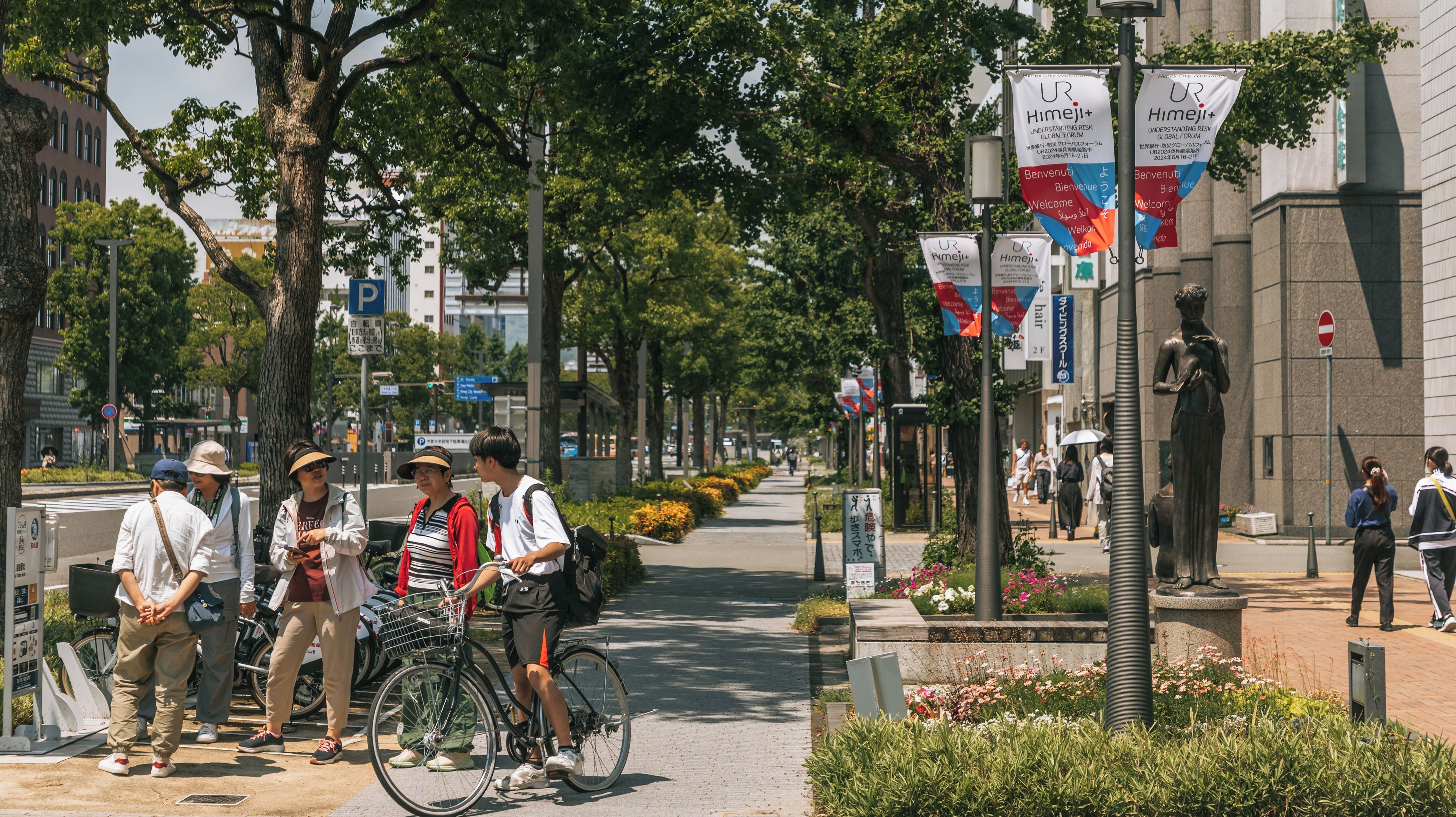 Rua arborizada em uma cidade, com uma estátua, pessoas caminhando e uma bicicleta estacionada na calçada.