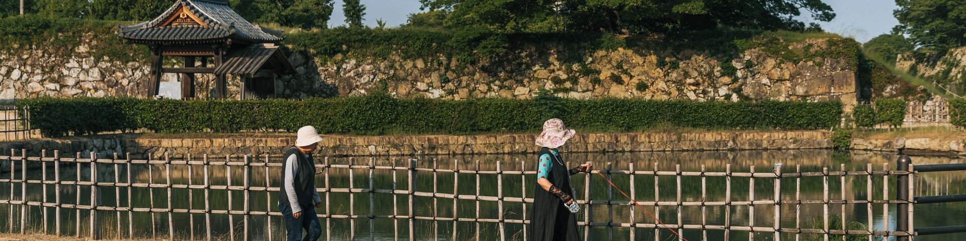 A traditional pavilion, a stone wall, and a wooden fence by a body of water.