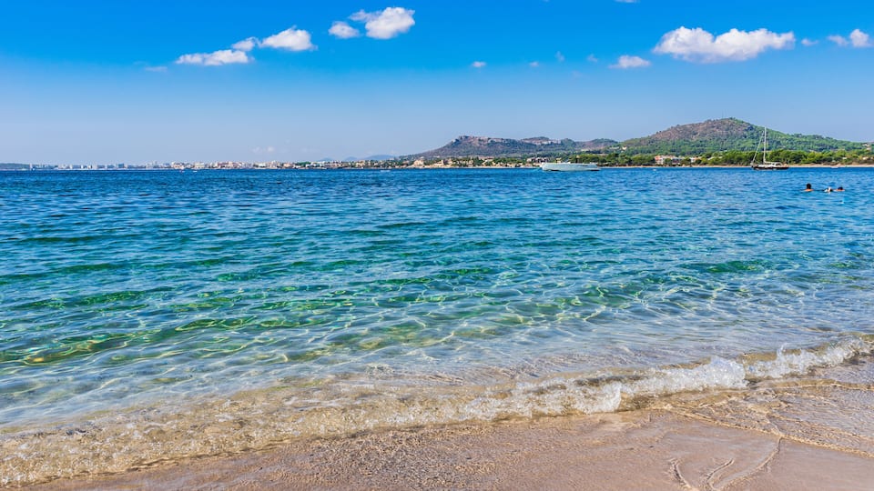 A clear blue sea with gentle waves, a sandy beach, and a distant mountain range.