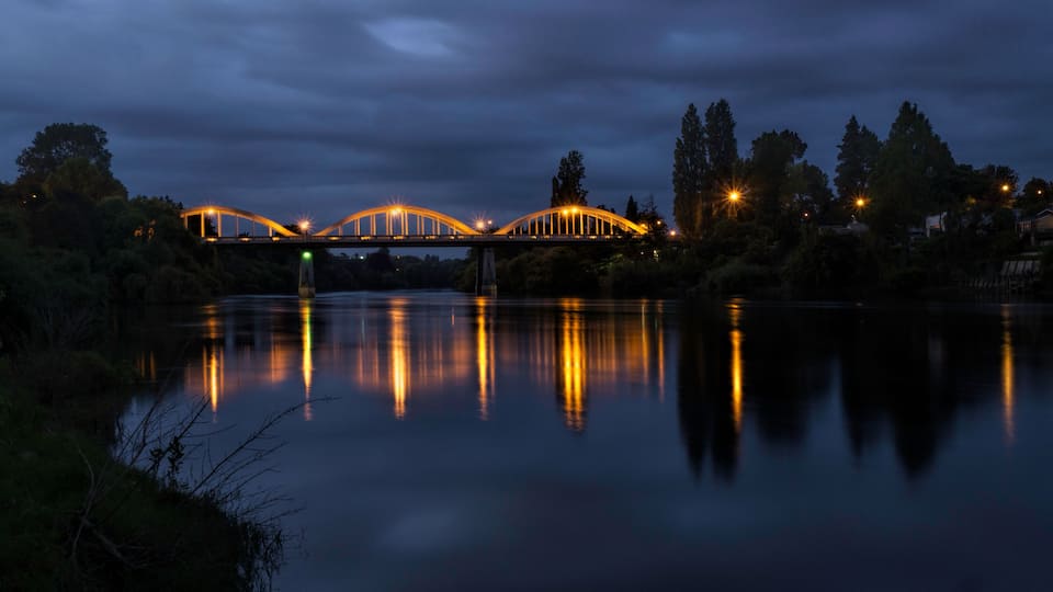 A bridge with lights reflecting on a calm river at night.