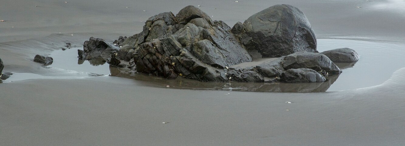 Una playa con grandes rocas y una cadena montañosa distante bajo cielos nublados.