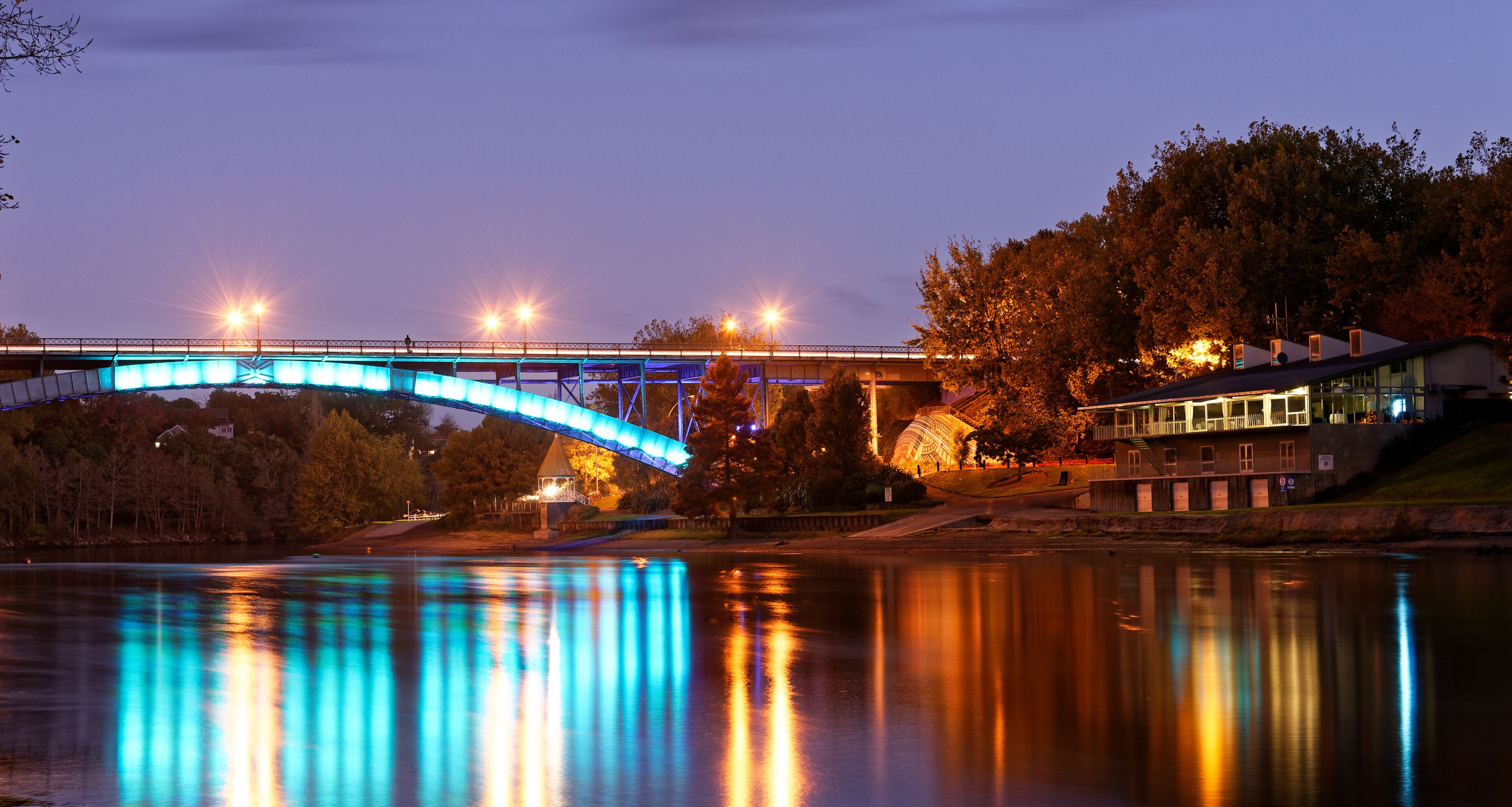 Un puente con luces azules reflejadas en el agua, rodeado de árboles y un edificio.