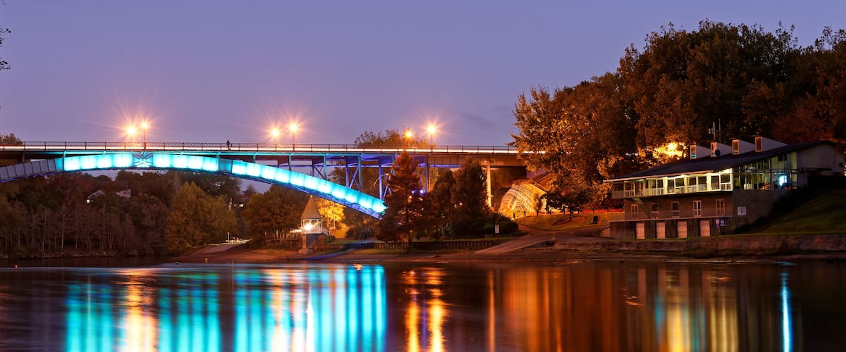 A bridge with blue lights reflecting on water, surrounded by trees and a building.