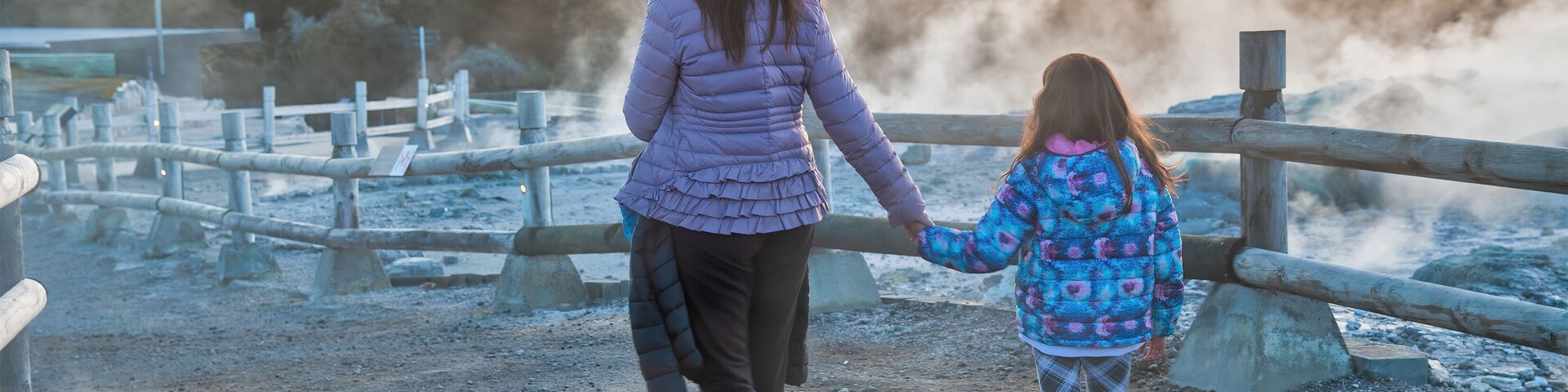 Uma mulher e uma criança caminhando sobre uma ponte de madeira sobre um corpo d'água, com vapor subindo da água.