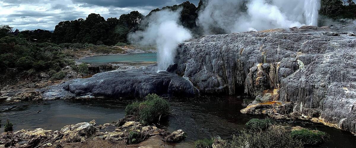 A geyser with water spouting from the ground, surrounded by rocky terrain and a cloudy sky.