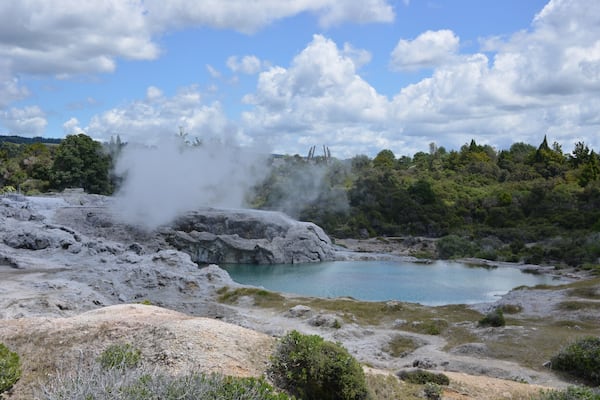 A geothermal area with a hot spring, steam rising, and a turquoise pool surrounded by rocky terrain and greenery.