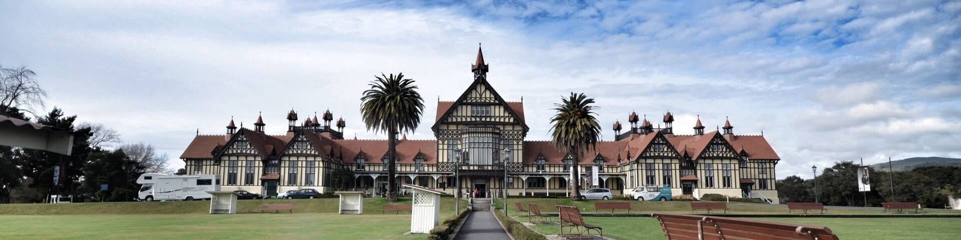 A large, historic building with a central tower and multiple smaller towers, surrounded by a well-maintained lawn and benches.