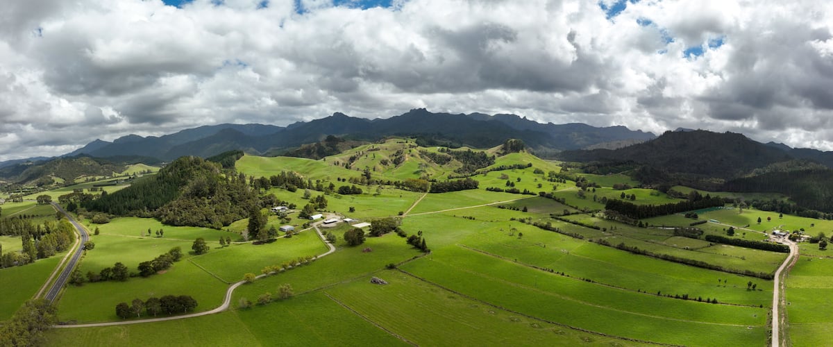 A lush green valley with rolling hills, a winding road, and distant mountains under a partly cloudy sky.