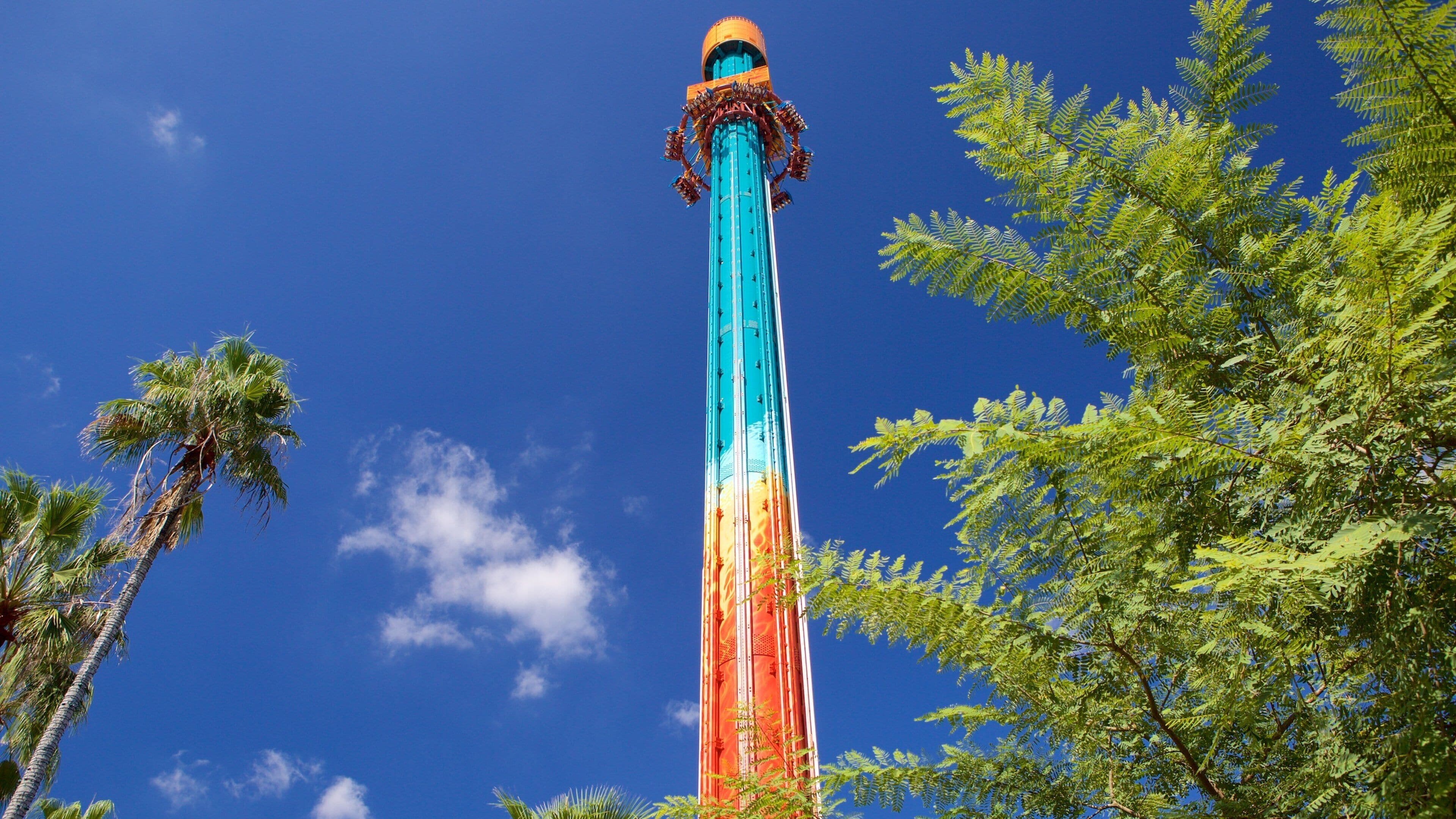 Een hoge, kleurrijke toren met een blauw, oranje en rood kleurverloop, omgeven door bomen en een helderblauwe lucht.