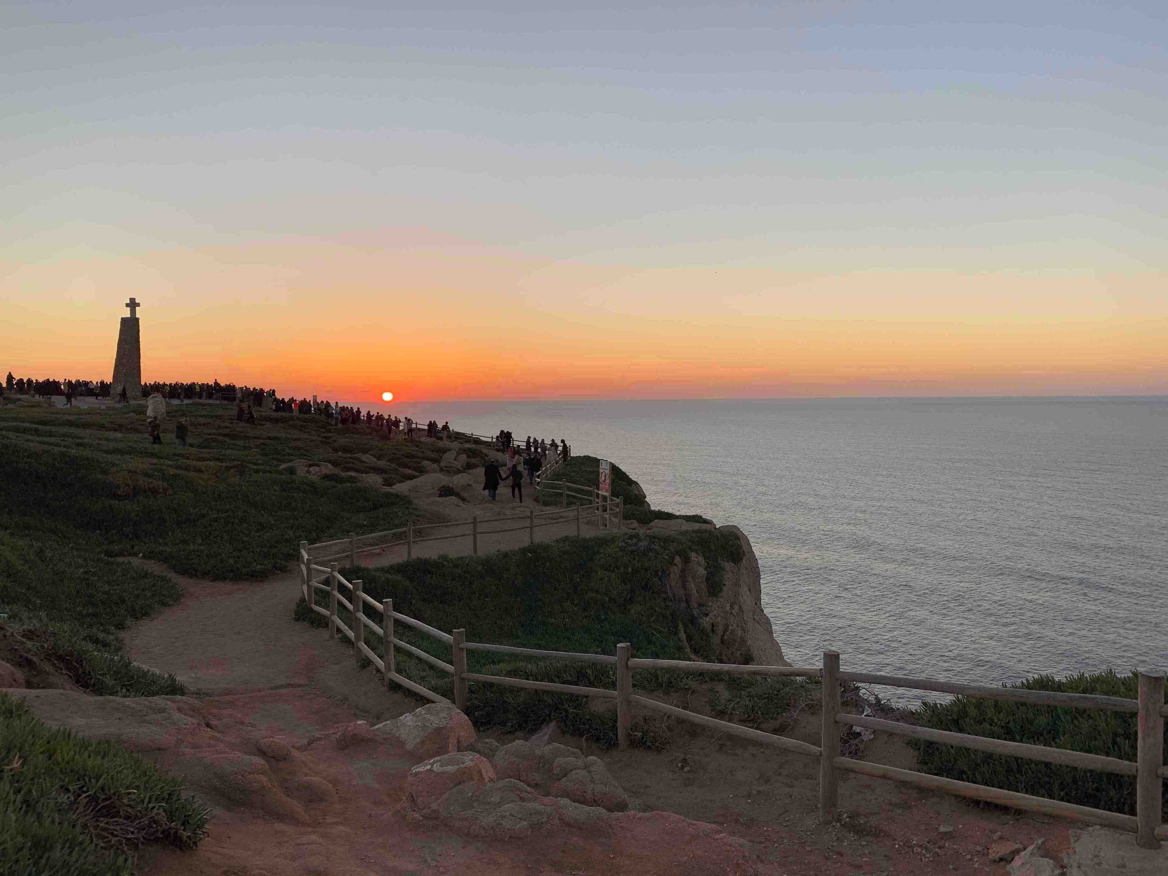 Vista del atardecer desde un sendero costero, con un faro y terreno rocoso.