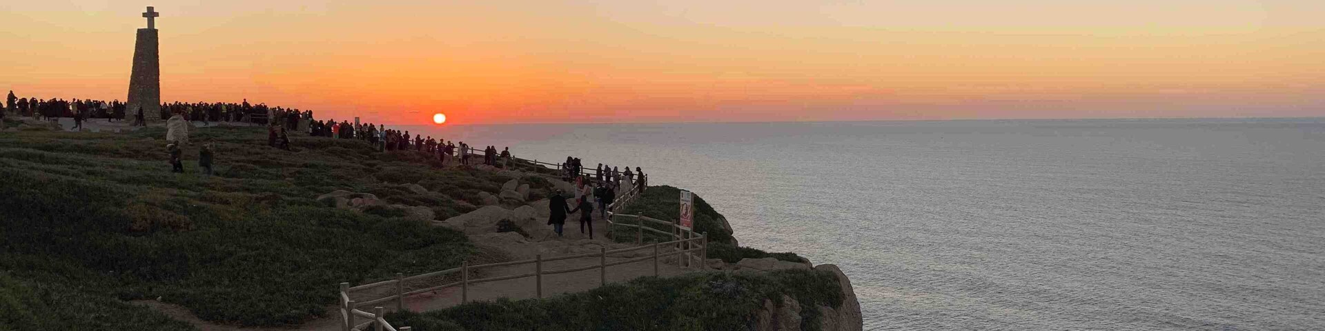 Vista del atardecer desde un sendero costero, con un faro y terreno rocoso.