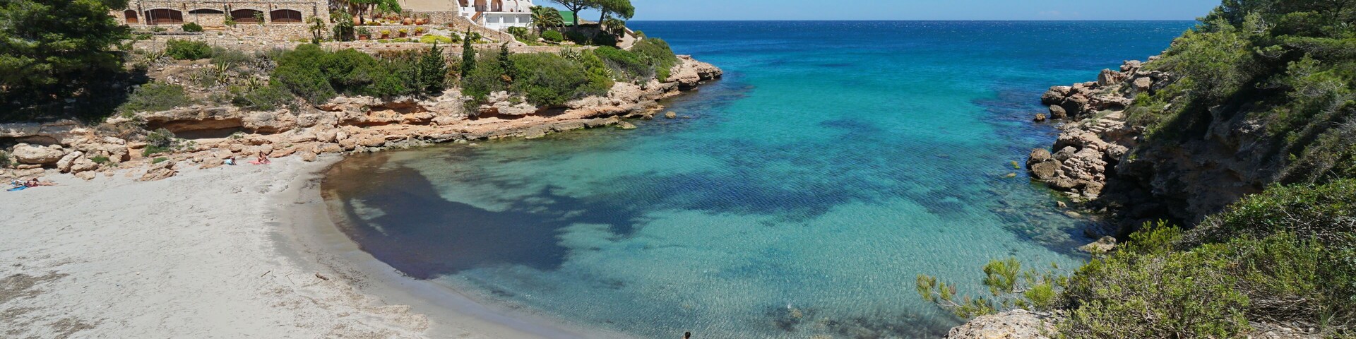 A coastal landscape with a clear blue sea, a sandy beach, rocky cliffs, and a white building with arches.
