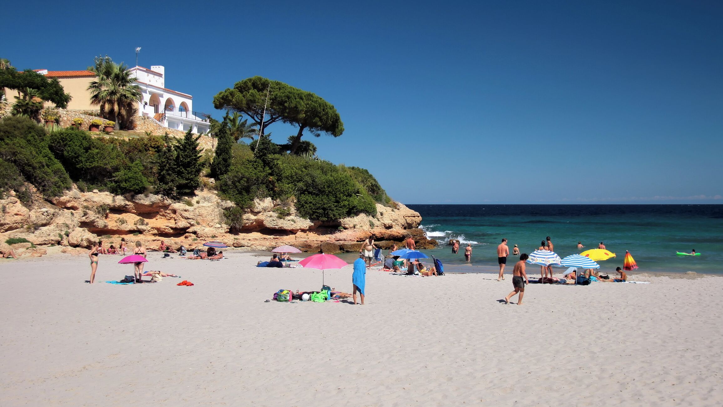 A sunny beach with people sunbathing and swimming, a white building with a red roof, and a rocky cliff with trees.