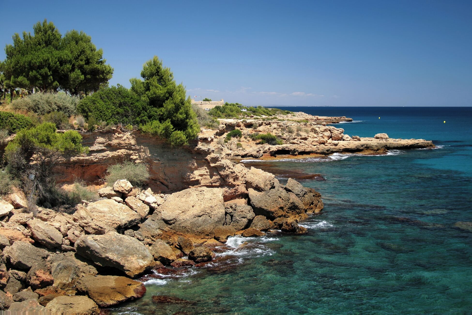 A rocky coastline with clear blue waters, green vegetation, and a distant building.