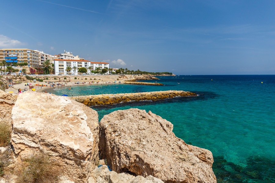 Uma vila costeira com águas turquesa cristalinas, formações rochosas e uma praia com pessoas e edifícios ao fundo.
