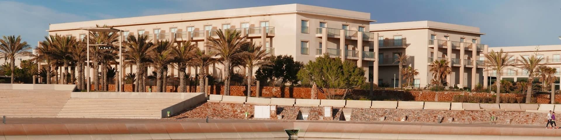 A modern building with a reflection in a calm body of water, surrounded by palm trees and a stone wall.
