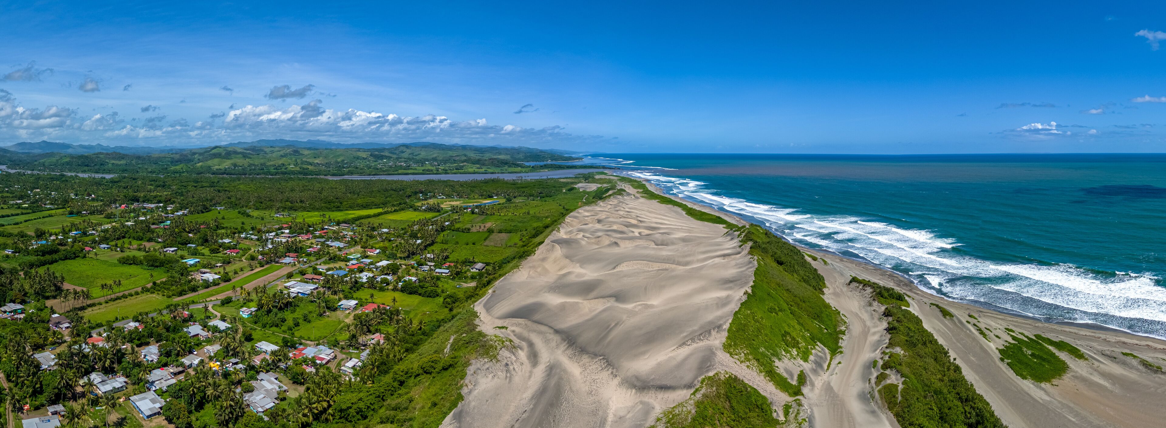 A drone panoramic aerial view of Fiji's Sigatoka Sand Dunes, showing the national park bordered by the Pacific Ocean, a local village, and lush green hills