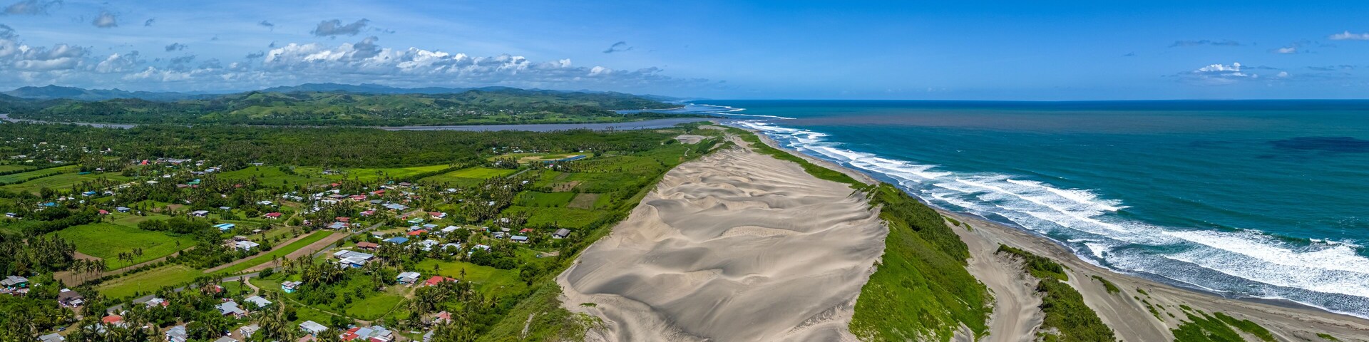 Un paisaje costero con playa de arena, un pequeño pueblo y colinas ondulantes al fondo.