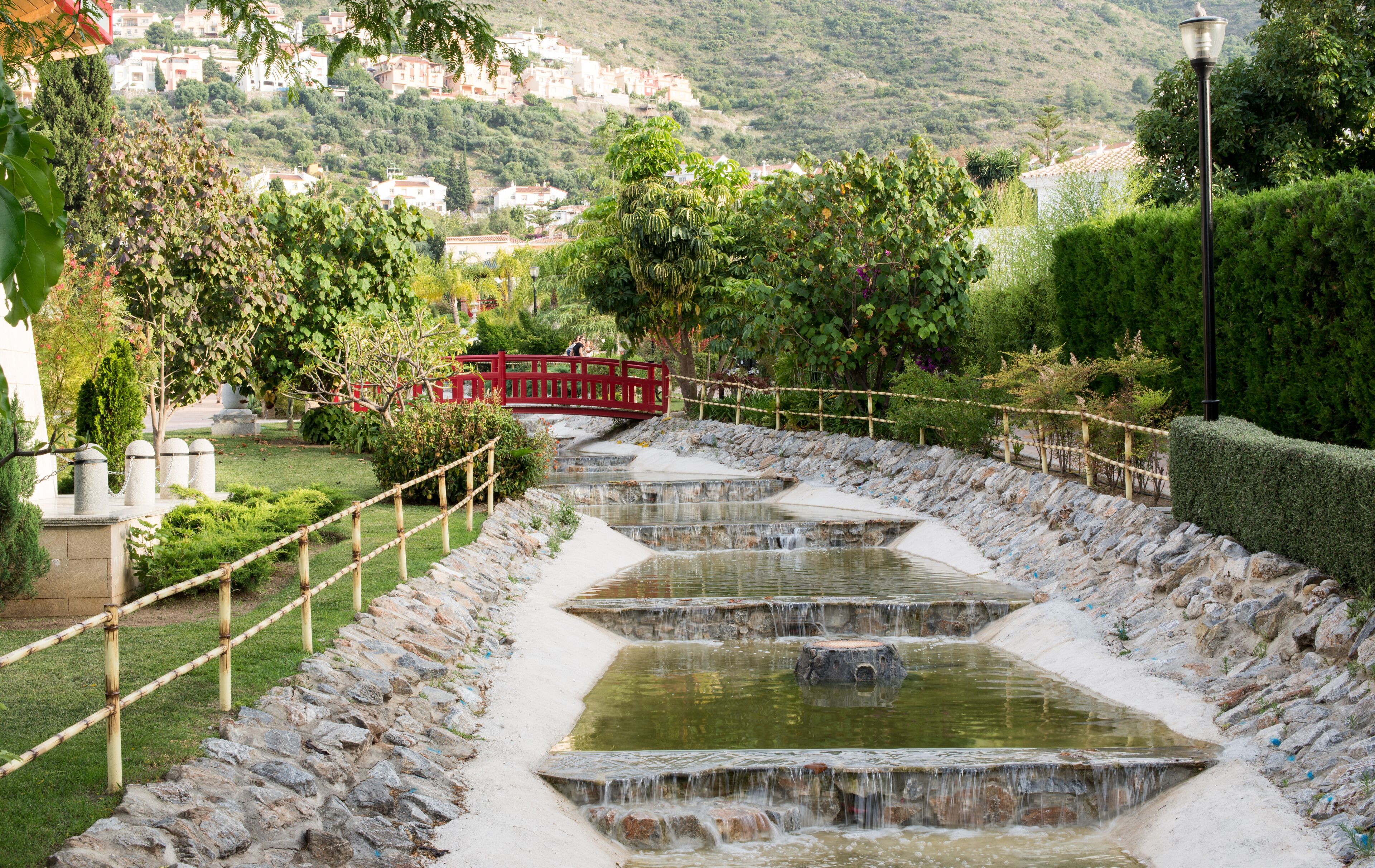 A terraced water feature with a red bridge, surrounded by greenery and a hillside in the background.