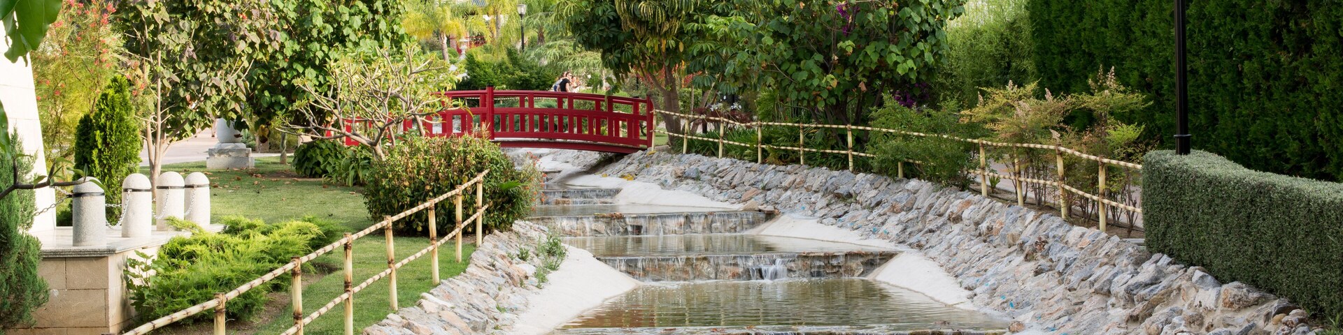 A terraced water feature with a red bridge, surrounded by greenery and a hillside in the background.
