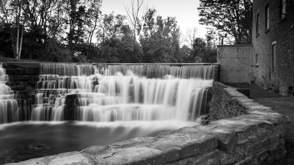 A black and white waterfall cascading over stone steps, surrounded by trees and a brick building.