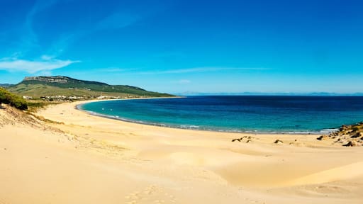 Un paisaje costero con una playa arenosa, mar azul cristalino y una colina con vegetación escasa.